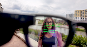 A woman stands behind a eyeglass window. The glass window shows closed captions.